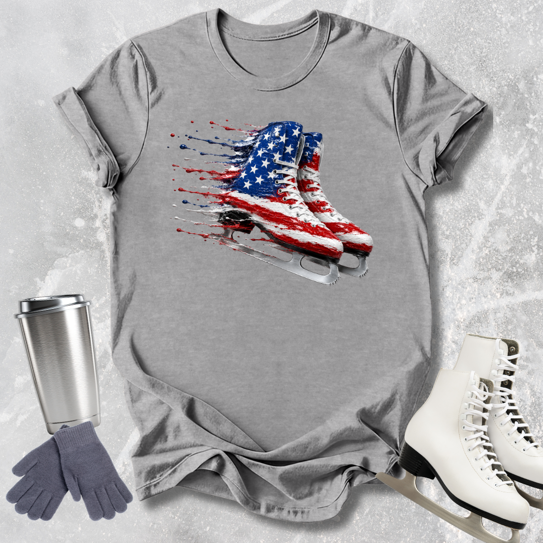 Gray t-shirt with American flag ice skate design, surrounded by a silver tumbler, gray gloves, and white ice skates on a textured gray background.