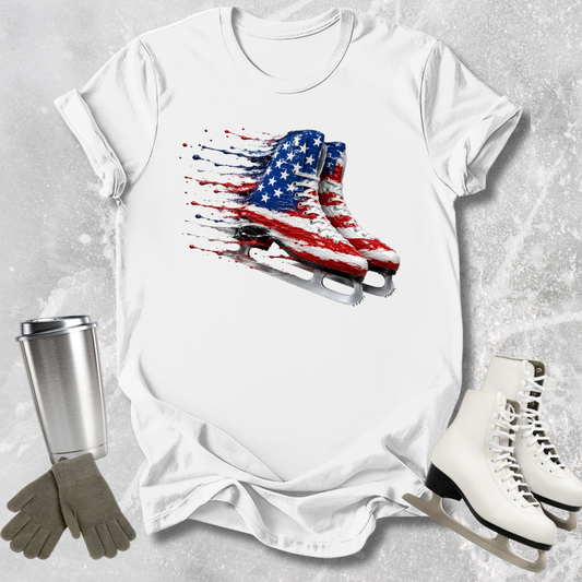 White t-shirt with American flag ice skate design, surrounded by ice skates and a tumbler on a textured surface.