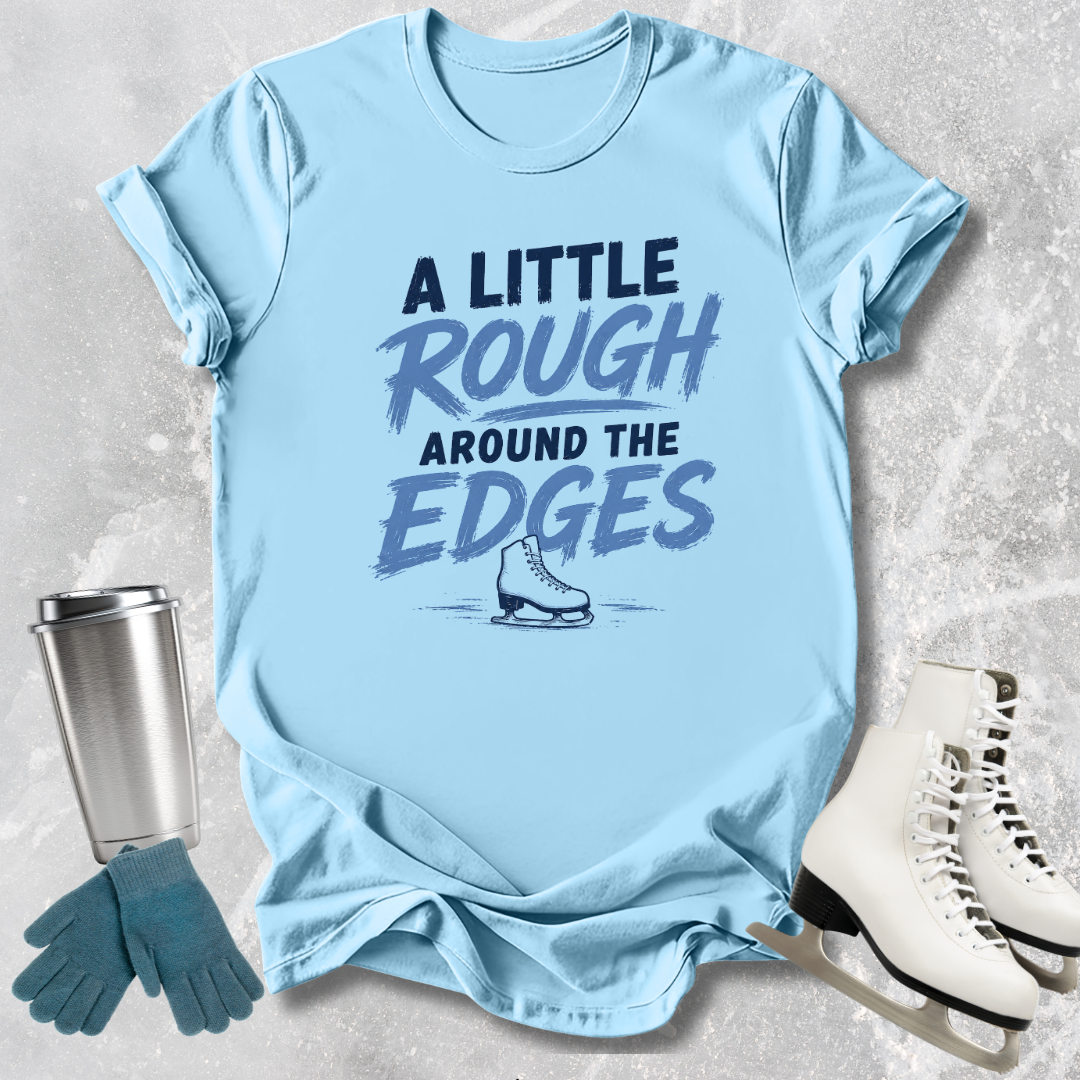 Light blue t-shirt with 'A Little Rough Around The Edges' text and graphic, next to a silver tumbler, green gloves, and white skates on a textured surface.