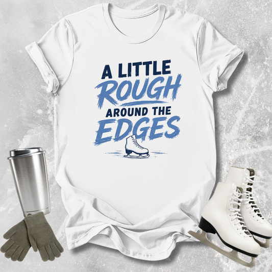 White t-shirt with 'A Little Rough Around The Edges' text and ice skate graphic, surrounded by ice skates, gloves, and a tumbler on a textured gray background.