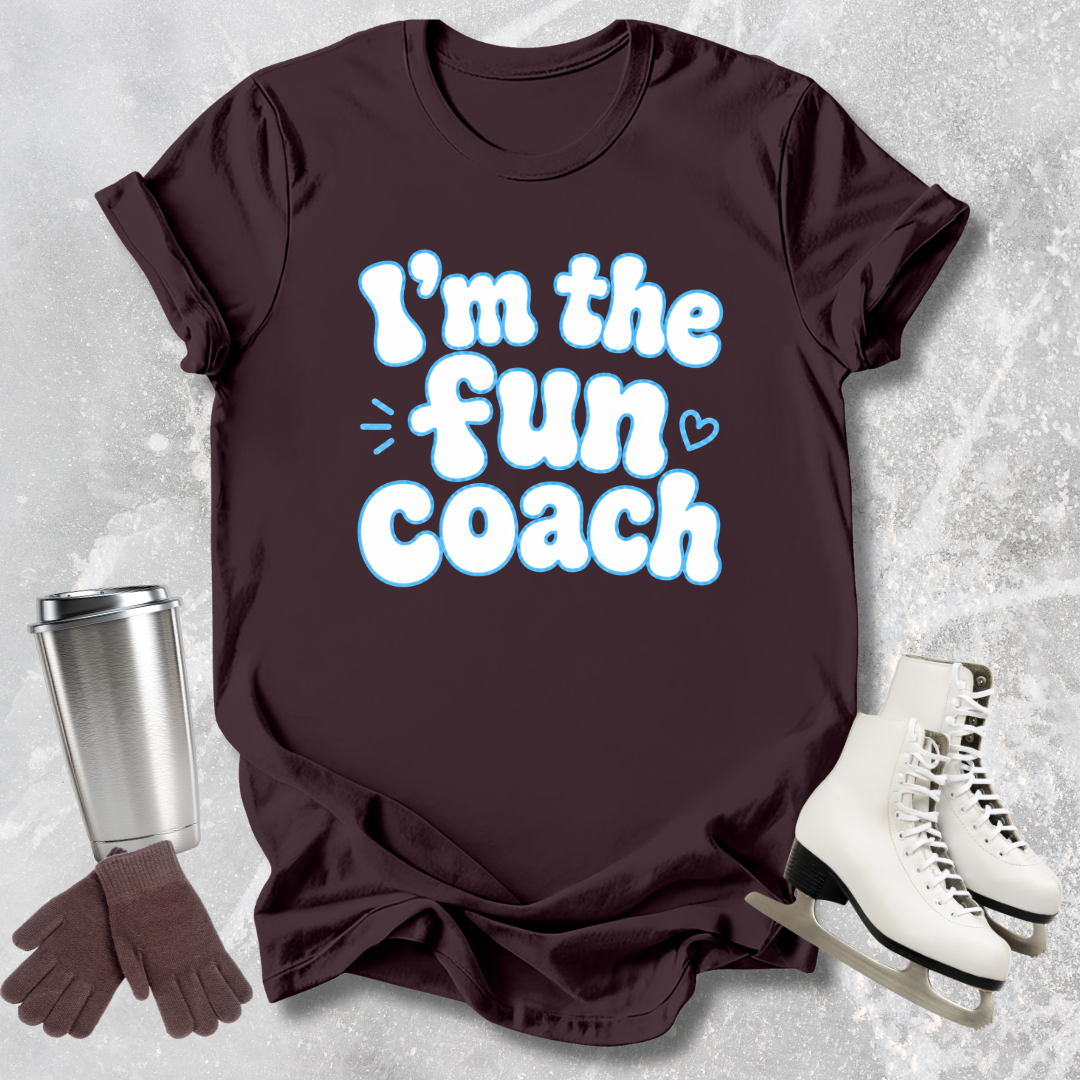 Brown t-shirt with 'I'm the fun coach' text, silver tumbler, brown gloves, and white skates on a textured gray background