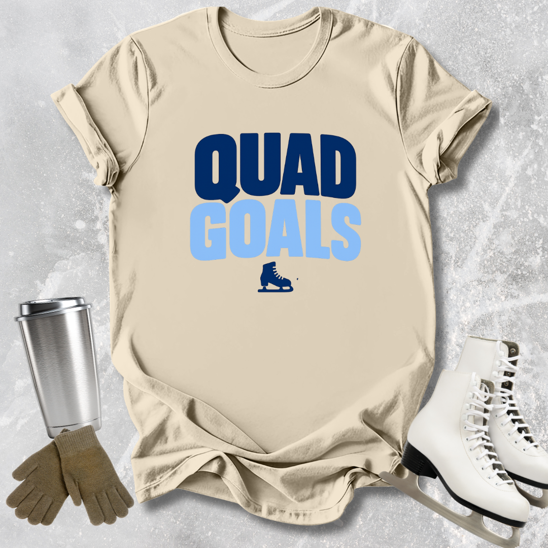 Beige t-shirt with 'Quad Goals' text and ice skates, next to a silver tumbler, brown gloves, and white ice skates on a light gray textured background.