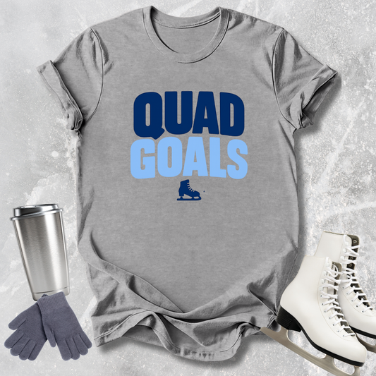 Gray t-shirt with 'Quad Goals' text, silver tumbler, gray gloves, and white skates on a textured surface.