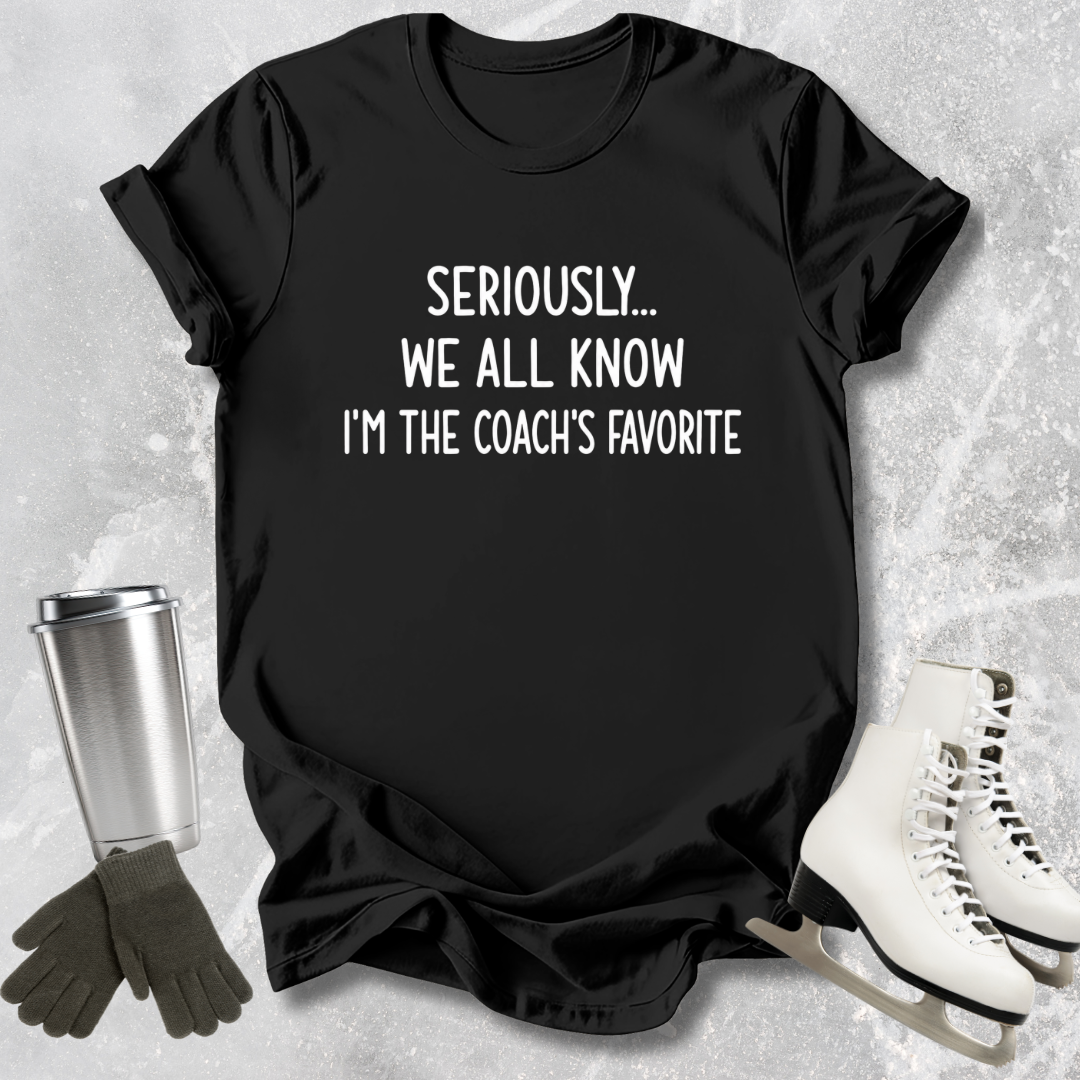 Black t-shirt with text 'Seriously... We all know I'm the coach's favorite' on a textured surface with accessories.