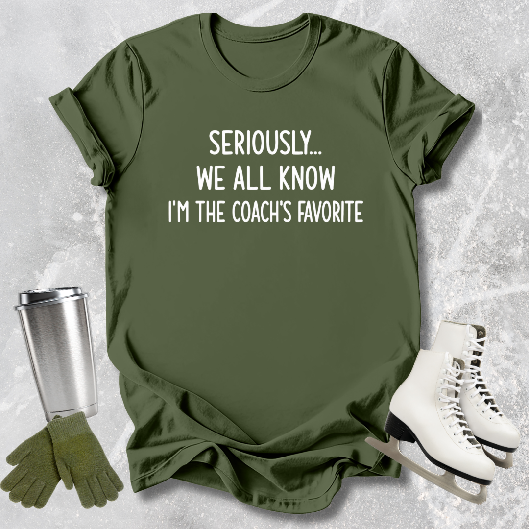 Green t-shirt with text 'Seriously... We all know I'm the coach's favorite' on a textured surface with accessories.