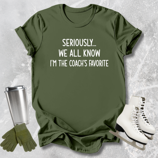 Green t-shirt with text 'Seriously... We all know I'm the coach's favorite' on a textured surface with accessories.