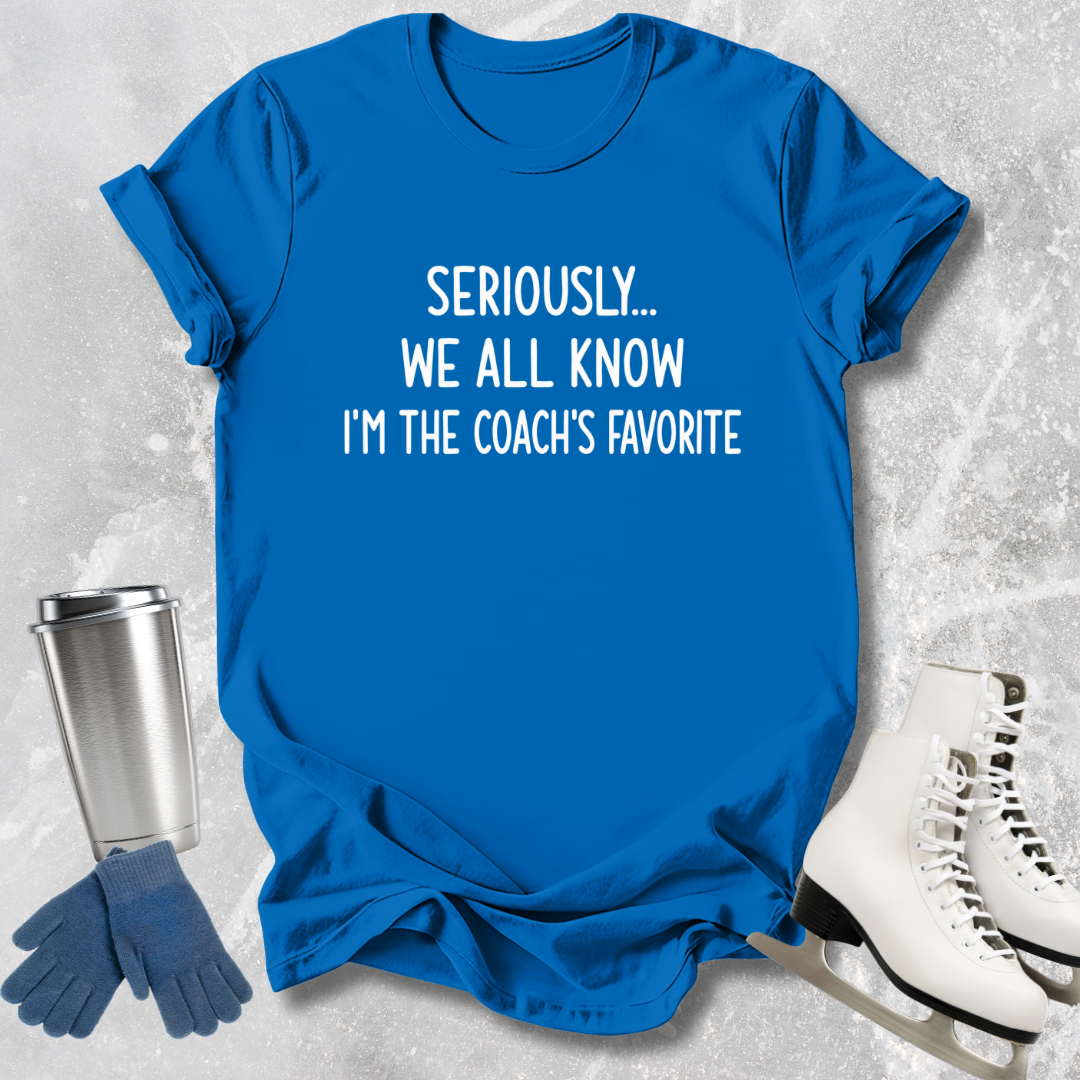 Blue t-shirt with text 'Seriously... We all know I'm the coach's favorite' on a textured surface with accessories.