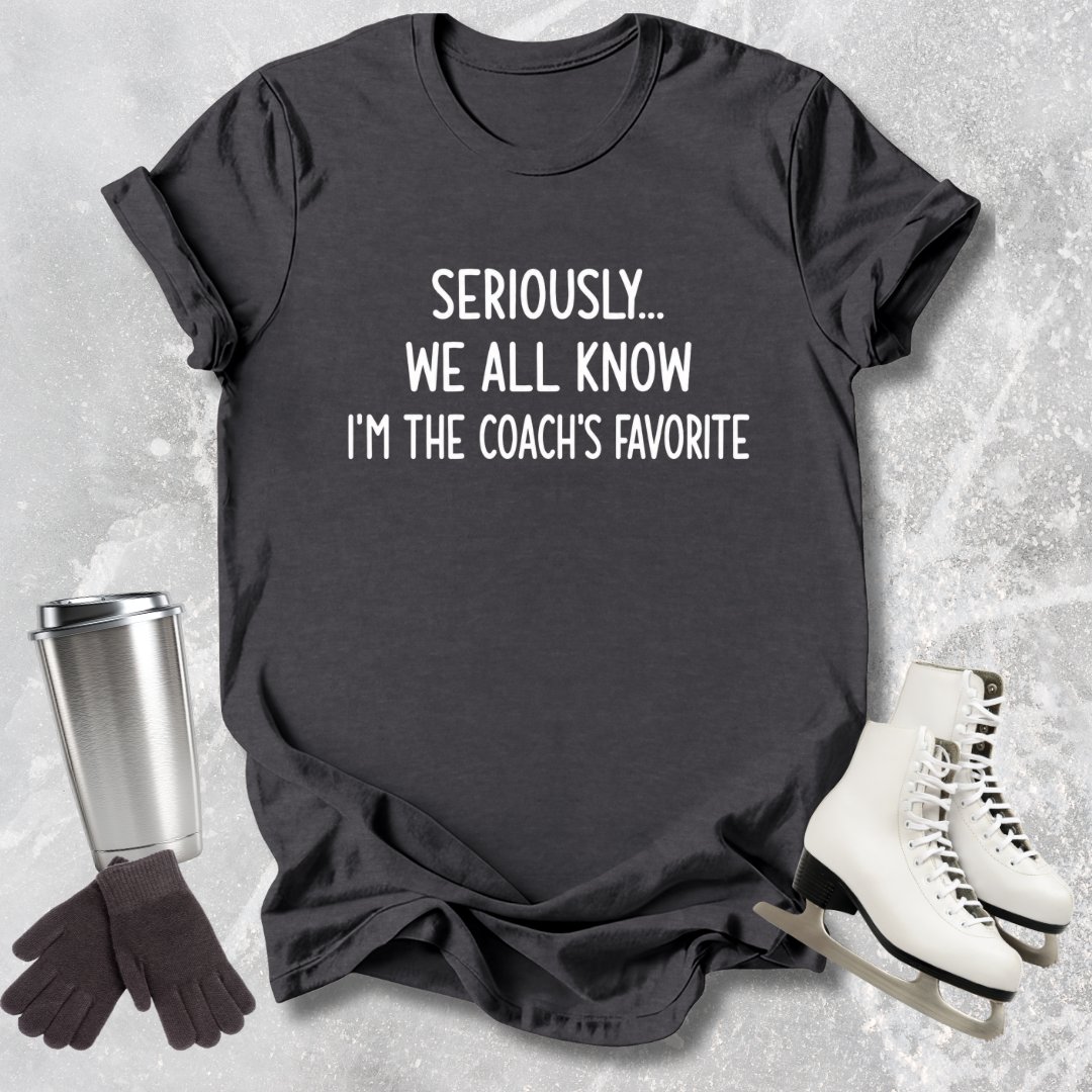 Black t-shirt with text 'Seriously... We all know I'm the coach's favorite' on a textured surface with accessories.