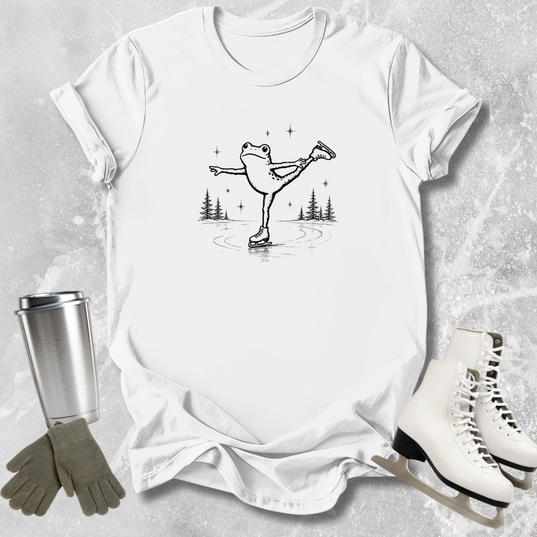 White t-shirt with a graphic of a frog on ice skates, surrounded by ice skates, gloves, and a tumbler on a textured surface.