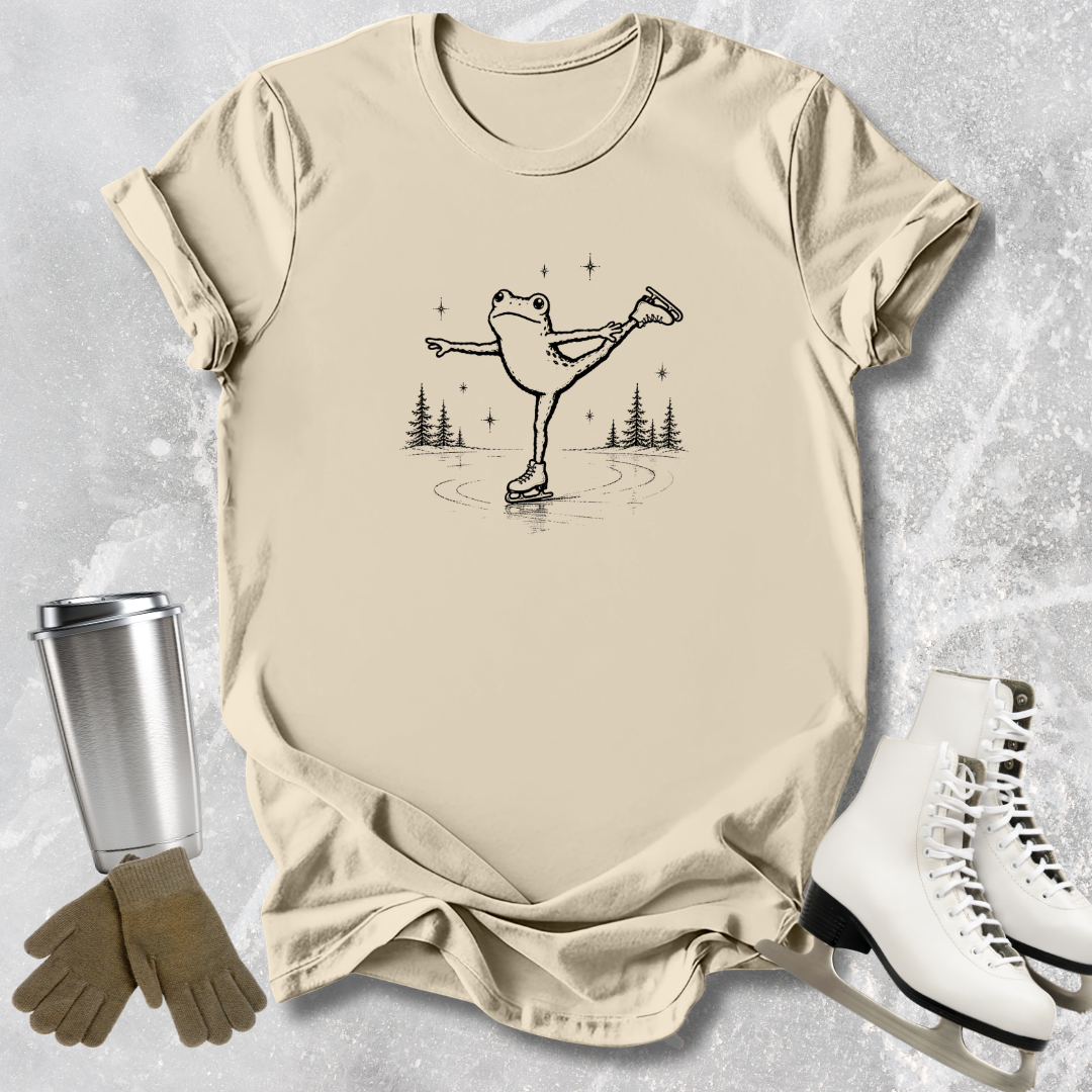 Beige t-shirt with a graphic of a frog on ice skates, next to a silver tumbler, brown gloves, and white ice skates on a textured gray background.