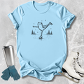 Light blue t-shirt with a frog ice skating design, next to a silver tumbler, green gloves, and white ice skates on a textured surface.