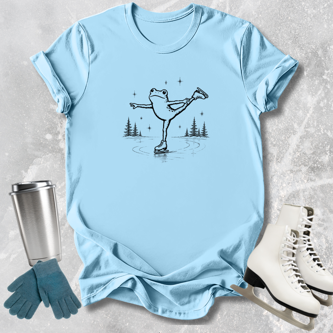 Light blue t-shirt with a frog ice skating design, next to a silver tumbler, green gloves, and white ice skates on a textured surface.