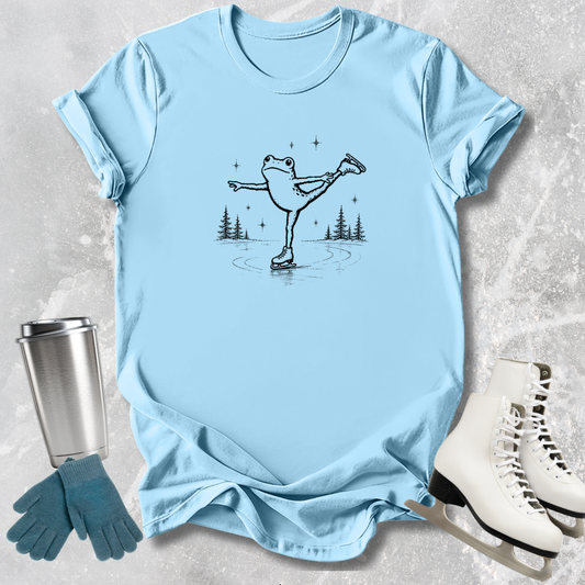 Light blue t-shirt with a frog ice skating design, next to a silver tumbler, green gloves, and white ice skates on a textured surface.