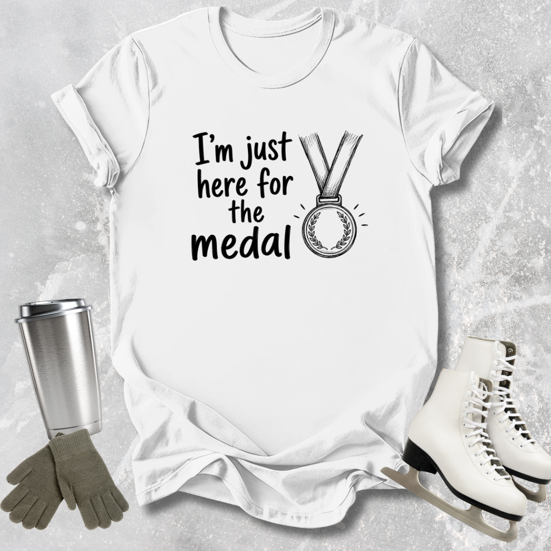 White t-shirt with 'I'm just here for the medal' text and graphic, surrounded by ice skates, gloves, and a tumbler on a light gray background.