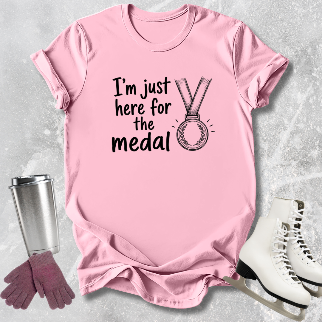 Pink t-shirt with 'I'm just here for the medal' text and graphic, next to a silver tumbler, purple gloves, and white skates on a light gray background.