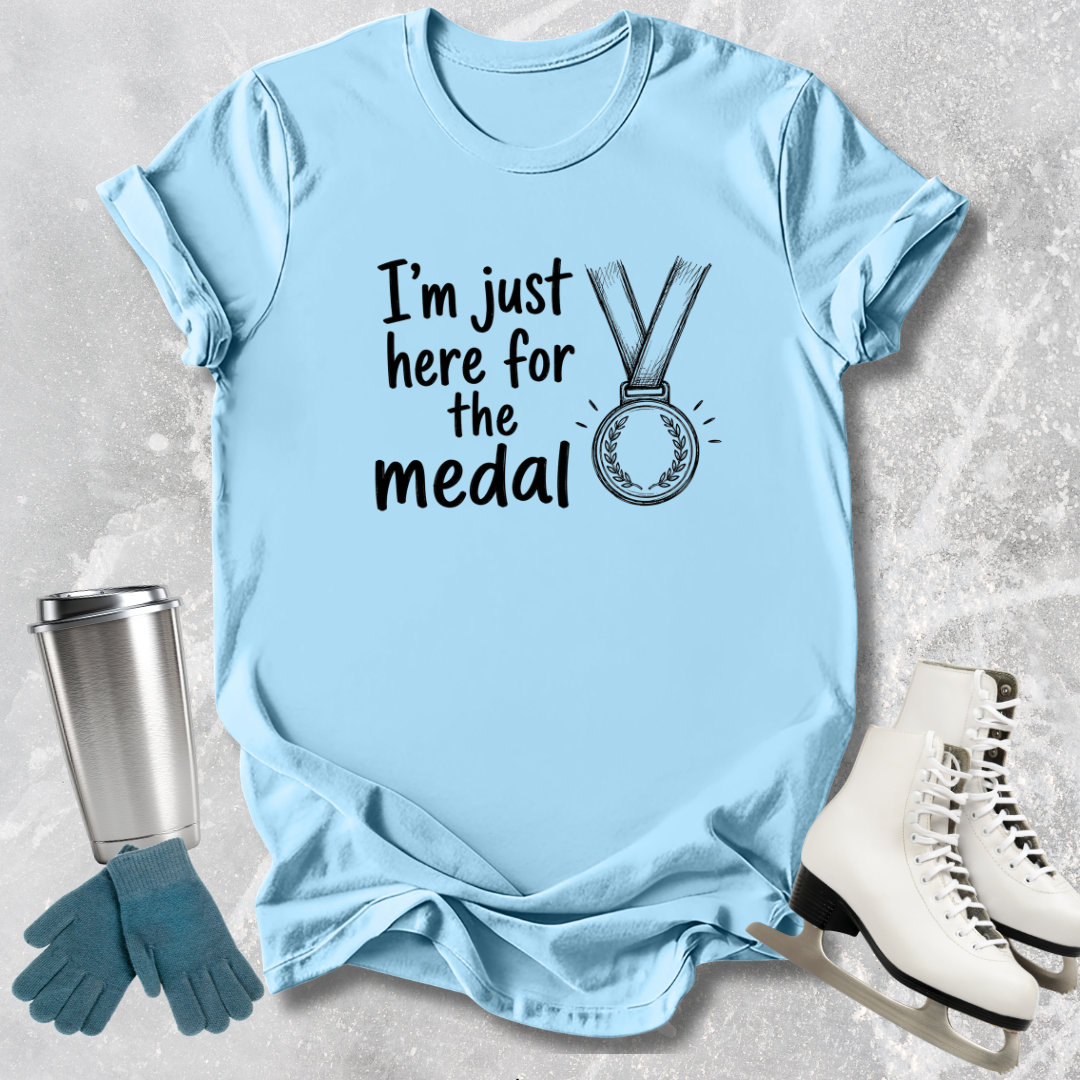 Light blue t-shirt with 'I'm just here for the medal' text and graphic, placed on a textured surface with accessories.