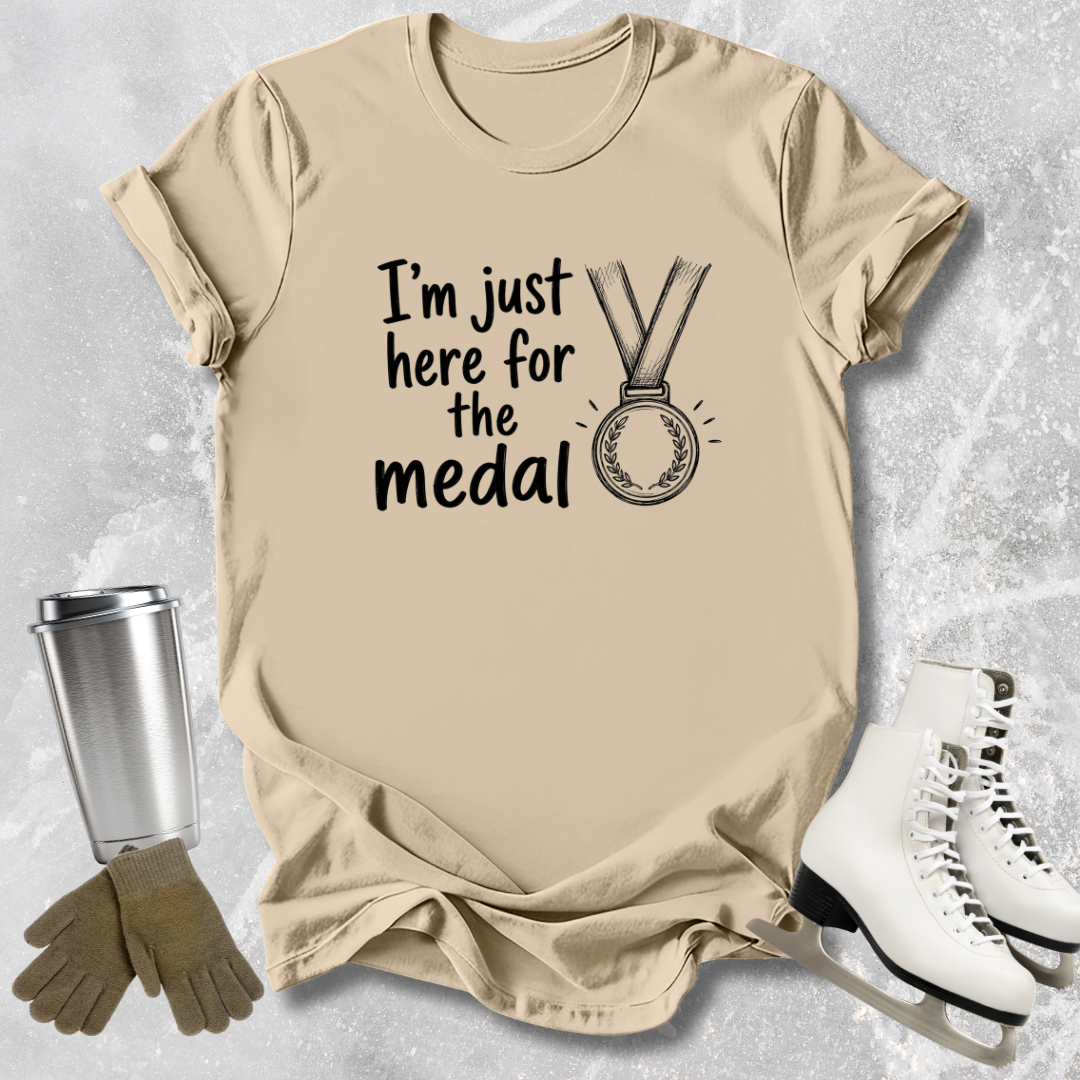 Beige t-shirt with 'I'm just here for the medal' text, next to ice skates and a tumbler on a textured surface.