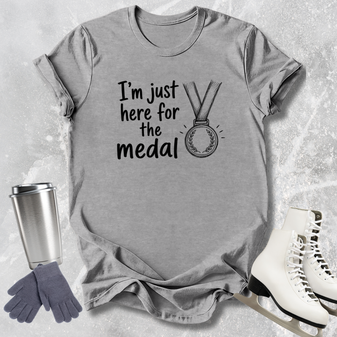 Gray t-shirt with 'I'm just here for the medal' text and graphic, next to a silver tumbler, gray gloves, and white skates on a textured surface.