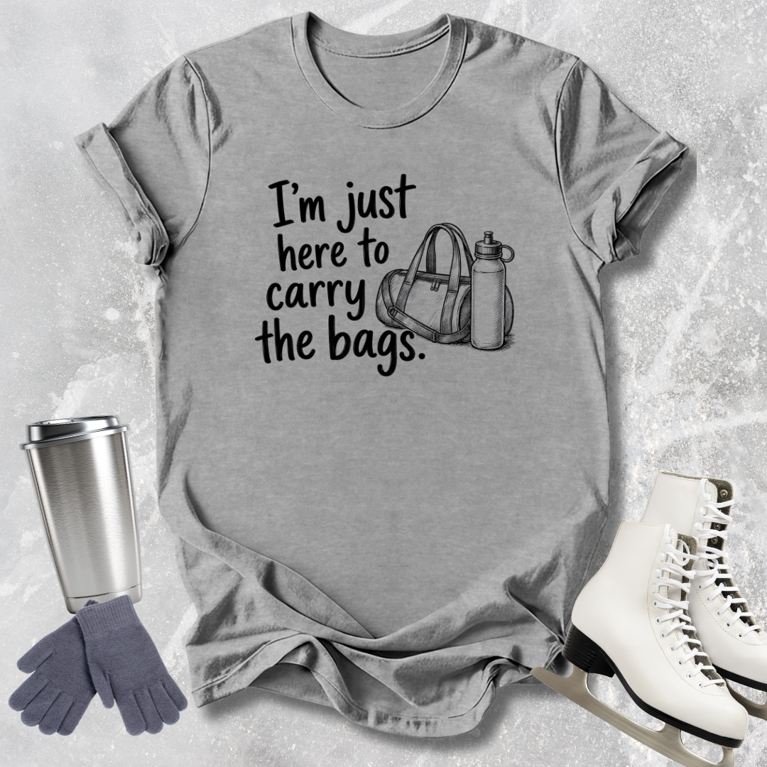 Gray t-shirt with text 'I'm just here to carry the bags' on a textured surface with accessories.