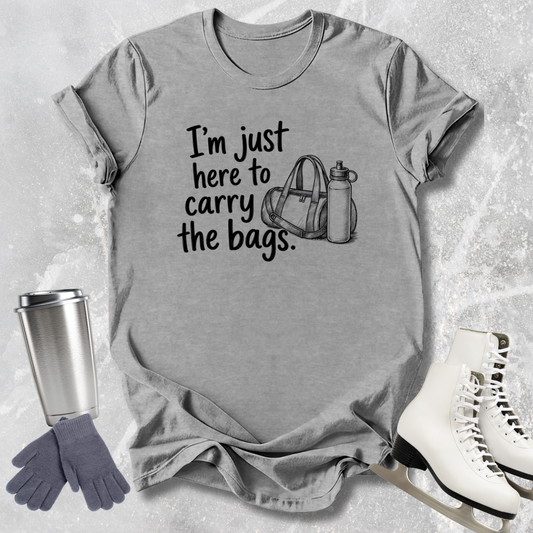 Gray t-shirt with text 'I'm just here to carry the bags' on a textured surface with accessories.