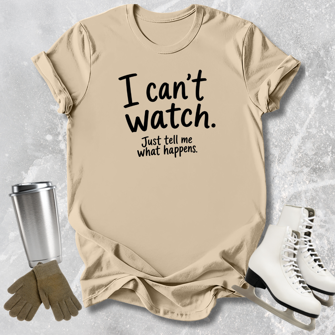 Beige t-shirt with text 'I can't watch. Just tell me what happens.' on a textured surface with a tumbler, gloves, and skates.