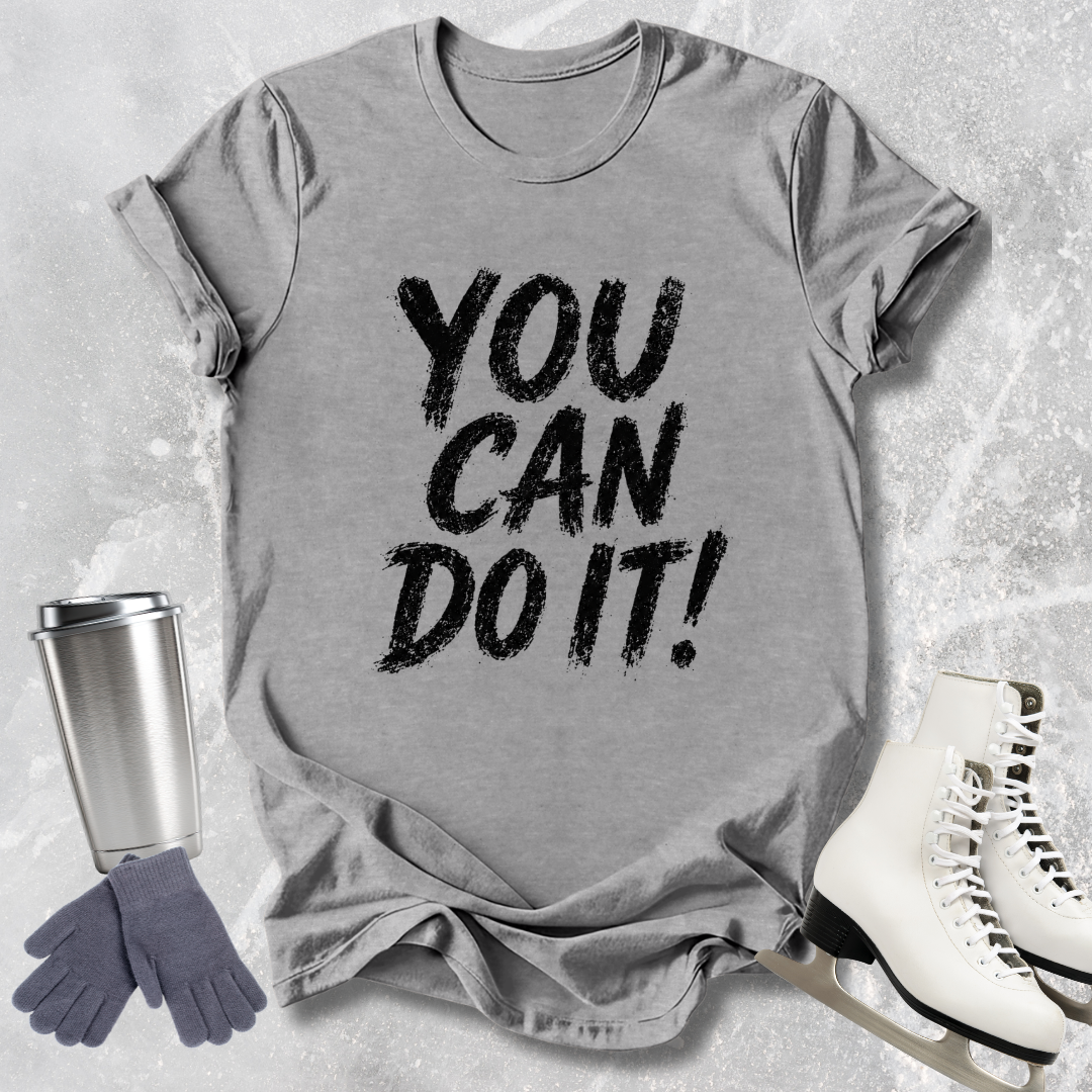 Gray t-shirt with 'You Can Do It!' text, next to a silver tumbler, gray gloves, and white skates on a textured surface.