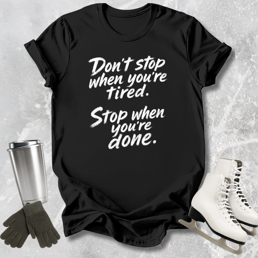 Black t-shirt with motivational quote, surrounded by a silver tumbler, black gloves, and white skates on a textured surface.