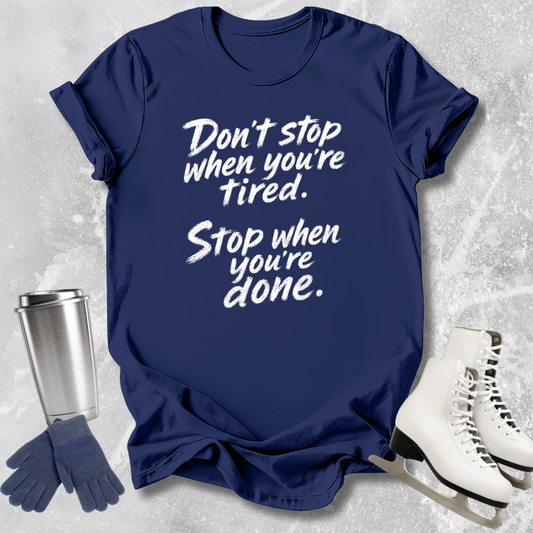 Navy blue t-shirt with motivational quote, next to a silver tumbler, gloves, and white skates on a textured gray background.