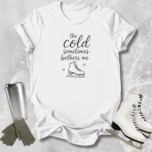 White t-shirt with 'the cold sometimes bothers me' text and ice skate graphic, surrounded by ice skates and a tumbler on a textured surface.