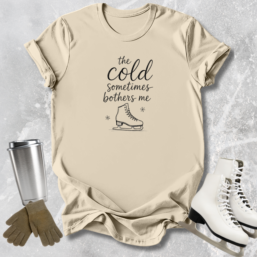 Beige t-shirt with text and graphic, next to a silver tumbler, brown gloves, and white skates on a textured surface.
