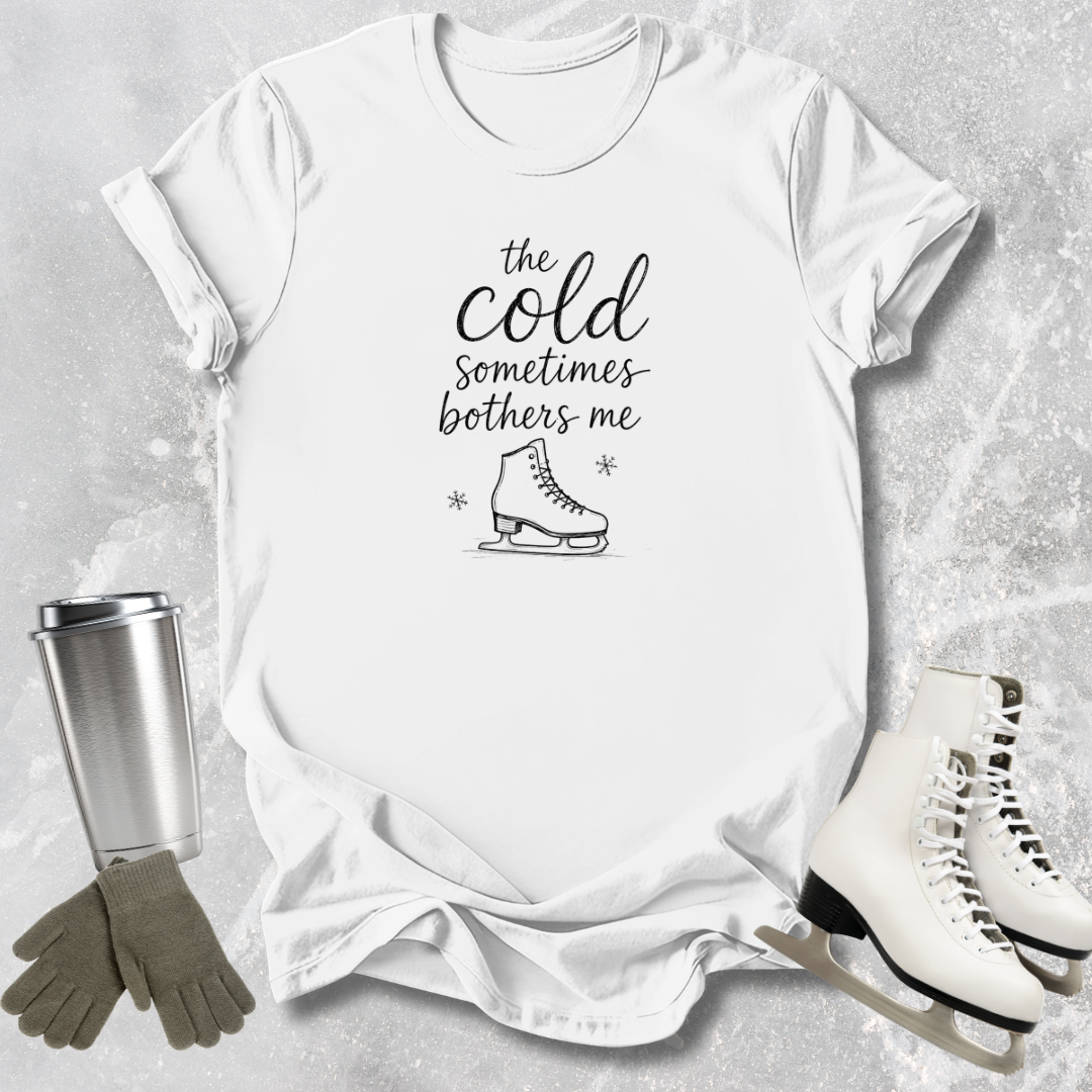 White t-shirt with 'the cold sometimes bothers me' text and ice skate graphic, surrounded by ice skates and a tumbler on a textured surface.