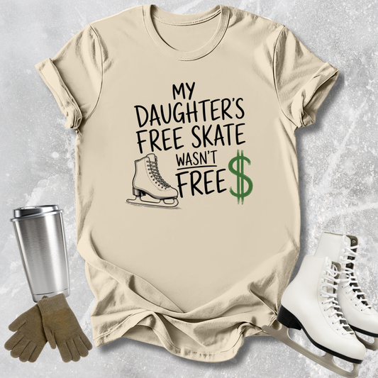 Beige t-shirt with text 'My daughter's free skate wasn't free' on a textured surface with ice skates and a tumbler.