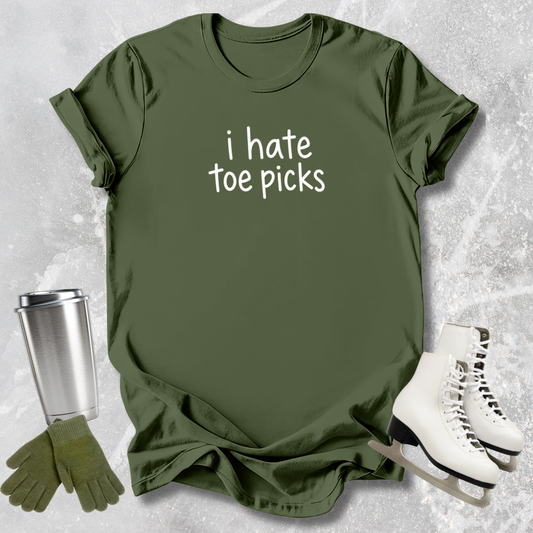 Green t-shirt with 'i hate toe picks' text, silver tumbler, blue gloves, and white ice skates on a textured gray background