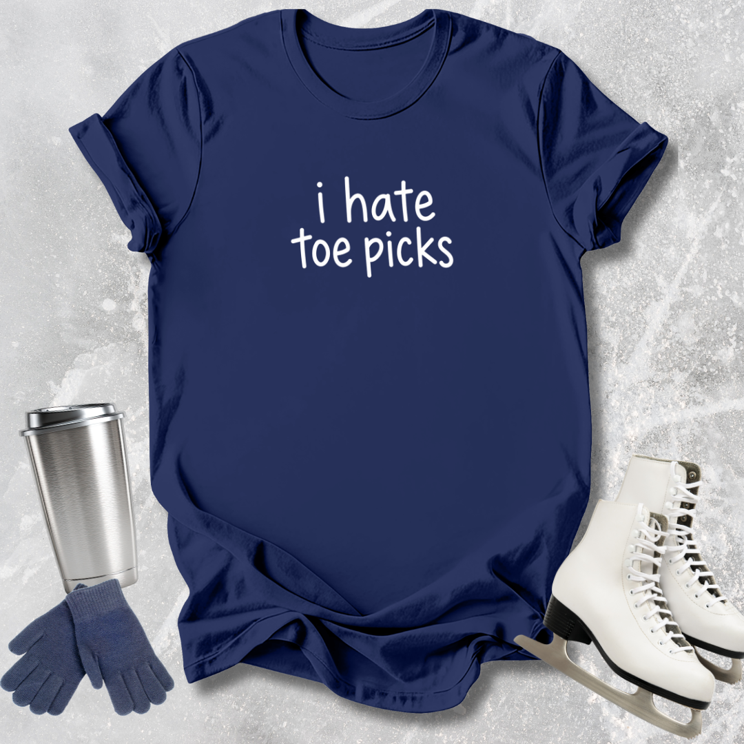 Navy blue t-shirt with 'i hate toe picks' text, next to a silver tumbler, blue gloves, and white ice skates on a textured gray background.