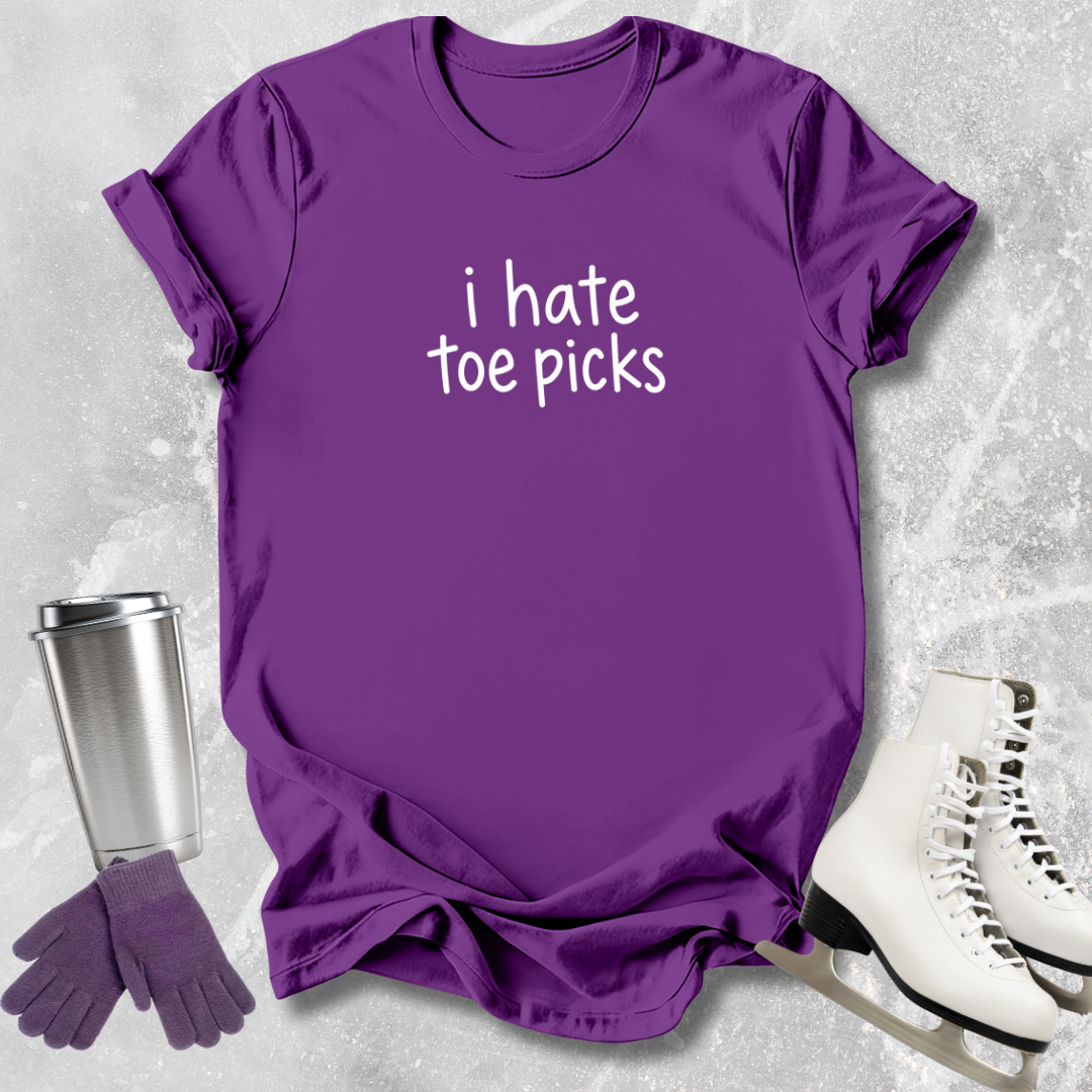 Purple t-shirt with 'i hate toe picks' text, next to a silver tumbler, purple gloves, and white ice skates on a textured gray background.
