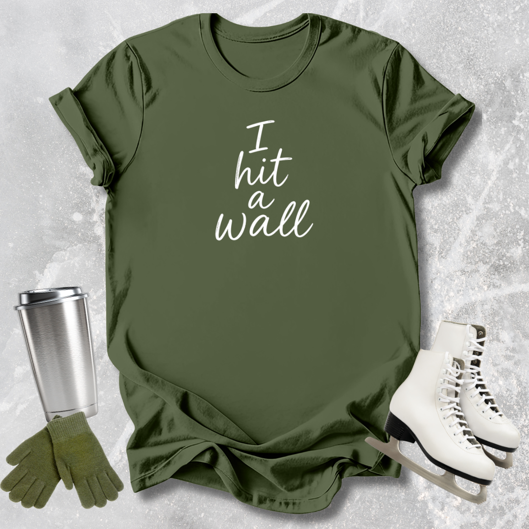Green t-shirt with 'I hit a wall' text, paired with a silver tumbler, green gloves, and white sneakers on a textured gray background.