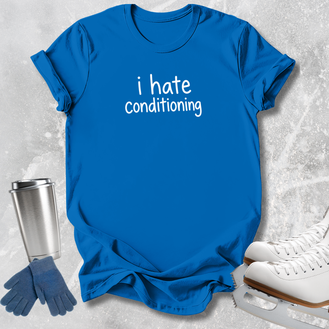 A bright blue short-sleeve T-shirt laid flat on a light gray textured surface, printed with the phrase 'i hate conditioning' in white lowercase text. Blue gloves, a silver travel tumbler, and white ice skates are arranged around it as styling props.