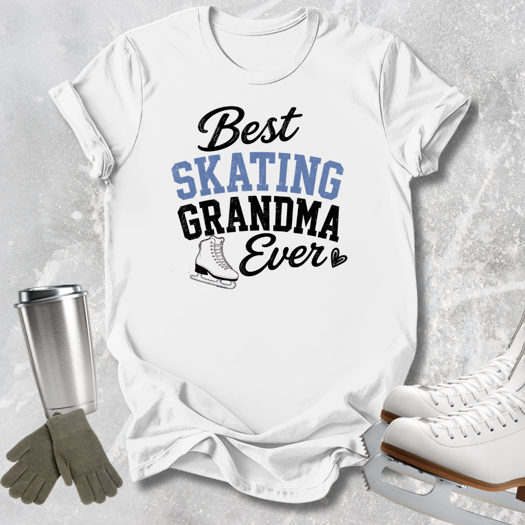 A white graphic T-shirt laid flat on a light gray textured surface, with the text 'Best SKATING GRANDMA Ever' in black and blue lettering and a small ice skate illustration. A stainless steel tumbler, gray gloves, and white ice skates are arranged around the shirt as props.