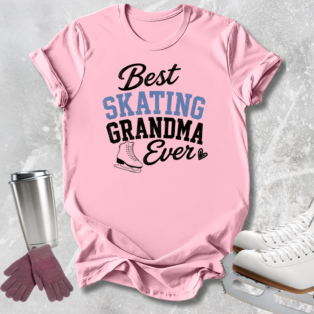 A pink short-sleeve graphic T-shirt laid flat on a light gray textured surface, with the text 'Best SKATING GRANDMA Ever' and a skater boot illustration. A stainless steel tumbler, pink gloves, and white ice skates are arranged around the shirt as styling props.