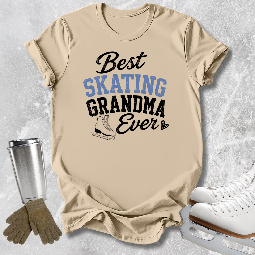 A beige graphic T-shirt laid flat on a light gray textured surface, with the words 'Best SKATING GRANDMA Ever' in black and light blue lettering and a small ice skate illustration. Ice skates, gloves, and a metal tumbler are arranged around the shirt as styling props.