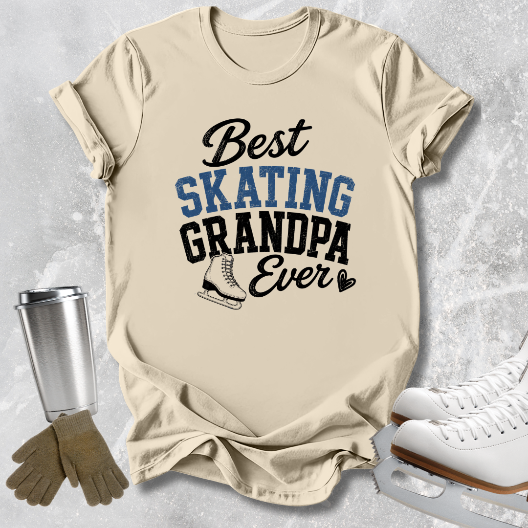 A beige graphic T-shirt laid flat on a light gray textured background, with the text 'Best SKATING GRANDPA Ever' in black and blue lettering and a small ice skate illustration. Surrounding props include white ice skates, tan gloves, and a silver tumbler.