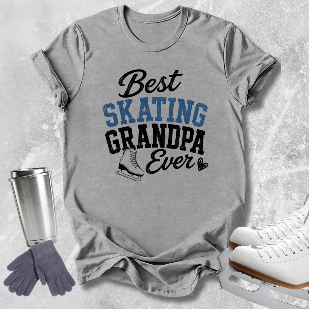 A heather gray graphic T-shirt laid flat on a light icy background, with the text 'Best SKATING GRANDPA Ever' and a small ice skate illustration. White ice skates, a silver travel tumbler, and gray gloves are arranged around it as styled props.
