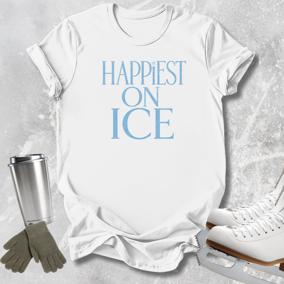 A white short-sleeve T-shirt laid flat on a light gray textured surface, printed with the phrase 'HAPPIEST ON ICE' in light blue. Ice skates, a metal tumbler, and gloves are arranged around it as styled props.