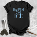 A black short-sleeve T-shirt laid flat on a light gray textured surface, printed with the phrase 'HAPPIEST ON ICE' in light blue. Ice skate boots, gloves, and a metal cup are arranged around the shirt as styled props.