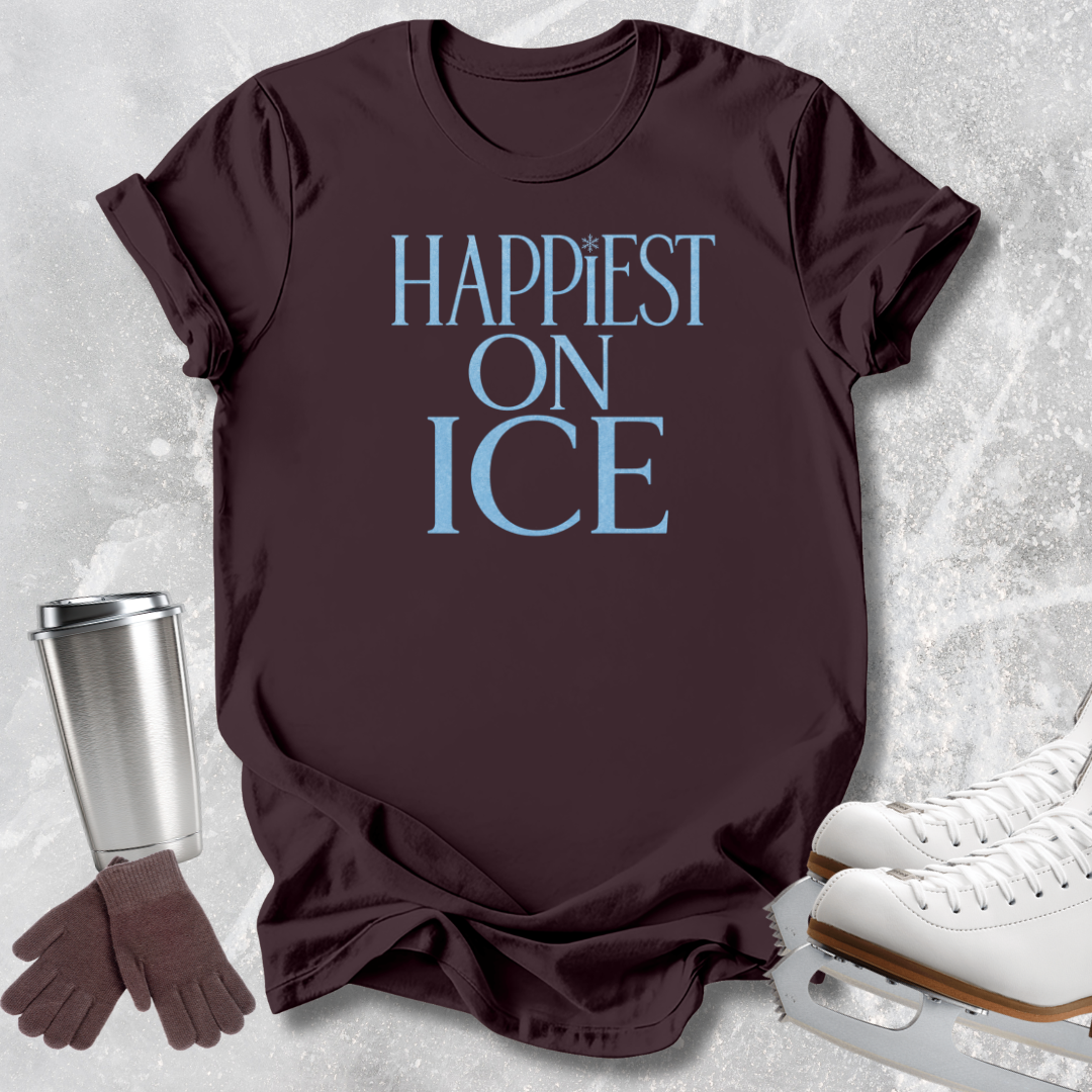 A dark brown short-sleeve t-shirt with the text 'HAPPIEST ON ICE' printed in light blue, styled in a flat-lay with ice skates, gloves, and a travel tumbler on a light gray textured background.