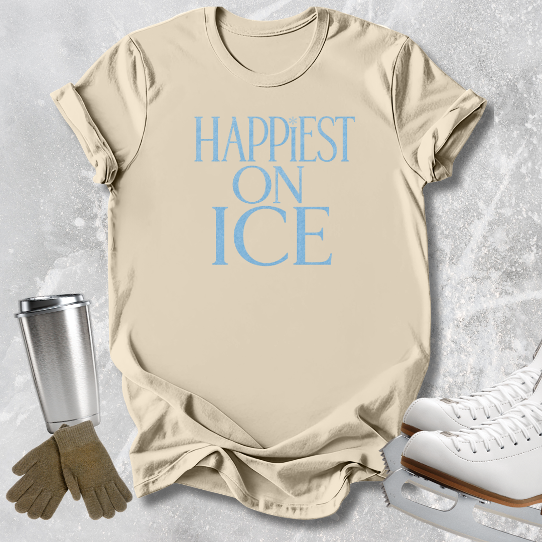 A beige short-sleeve T-shirt with the phrase 'HAPPIEST ON ICE' printed in light blue across the chest, styled in a flat lay with ice skates, gloves, and a travel tumbler on a light gray textured background.