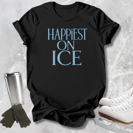 A black short-sleeve T-shirt laid flat on a light gray textured surface, printed with the phrase 'HAPPIEST ON ICE' in light blue. Ice skate boots, gloves, and a metal cup are arranged around the shirt as styled props.
