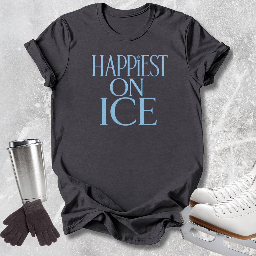 A dark charcoal gray graphic t-shirt with the text 'HAPPIEST ON ICE' printed in light blue. The t-shirt is styled in a flat-lay arrangement on a light gray textured background, accompanied by ice skates, a stainless steel travel mug, and a pair of black gloves.