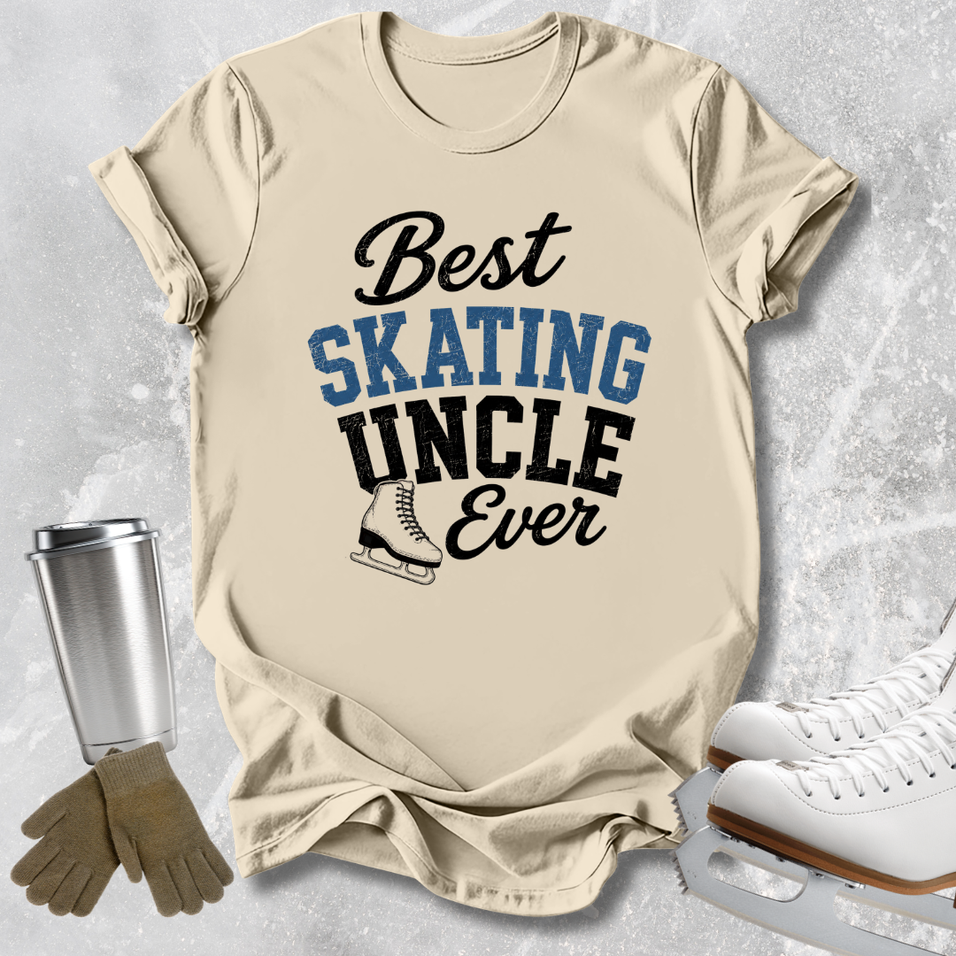 A beige graphic T-shirt laid flat on a light gray textured surface, with the text 'Best SKATING UNCLE Ever' in black and blue lettering and a small ice skate illustration. Surrounding props include a silver tumbler, tan gloves, and white ice skates.
