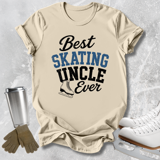 A beige graphic T-shirt laid flat on a light gray textured surface, with the text 'Best SKATING UNCLE Ever' in black and blue lettering and a small ice skate illustration. Surrounding props include a silver tumbler, tan gloves, and white ice skates.